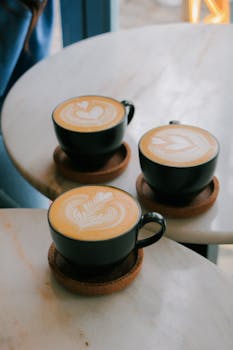 Three cappuccinos with intricate latte art on round white marble tables in a cafe.