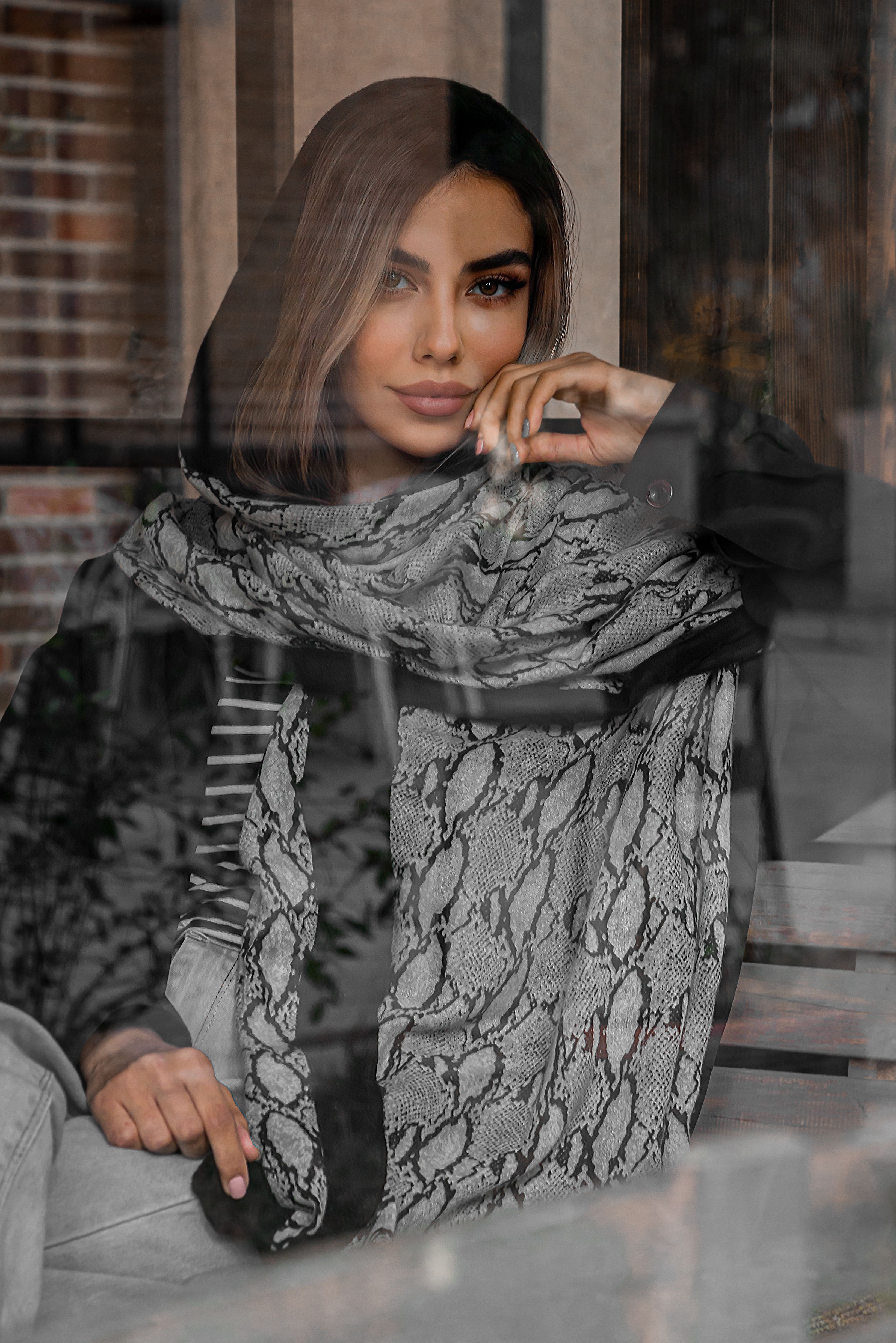 Stylish woman with patterned scarf posing behind a glass window indoors.