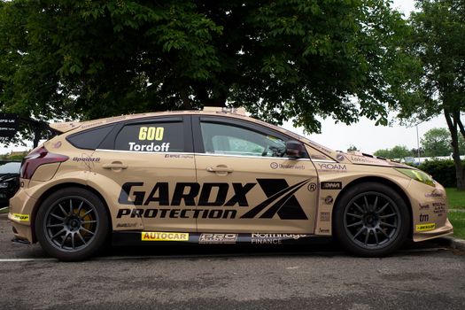 A side view of a racing car with promotional decals, parked under a green tree.