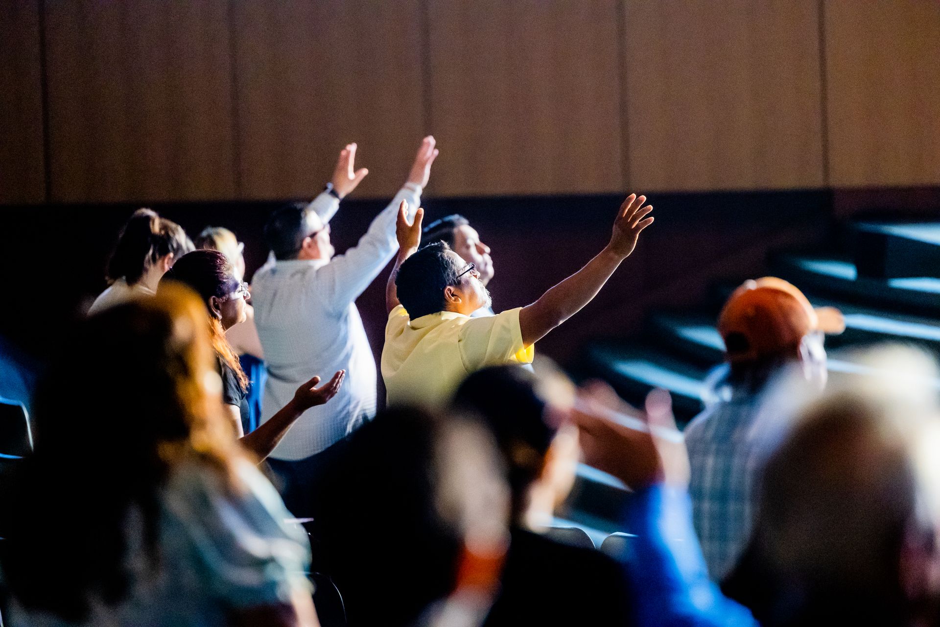 A diverse crowd engaged in worship indoors, raising hands in collective faith and devotion.