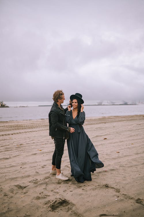 Woman in Black Dress with Hat Standing Beside a Man in Black Jacket on Beach Sand