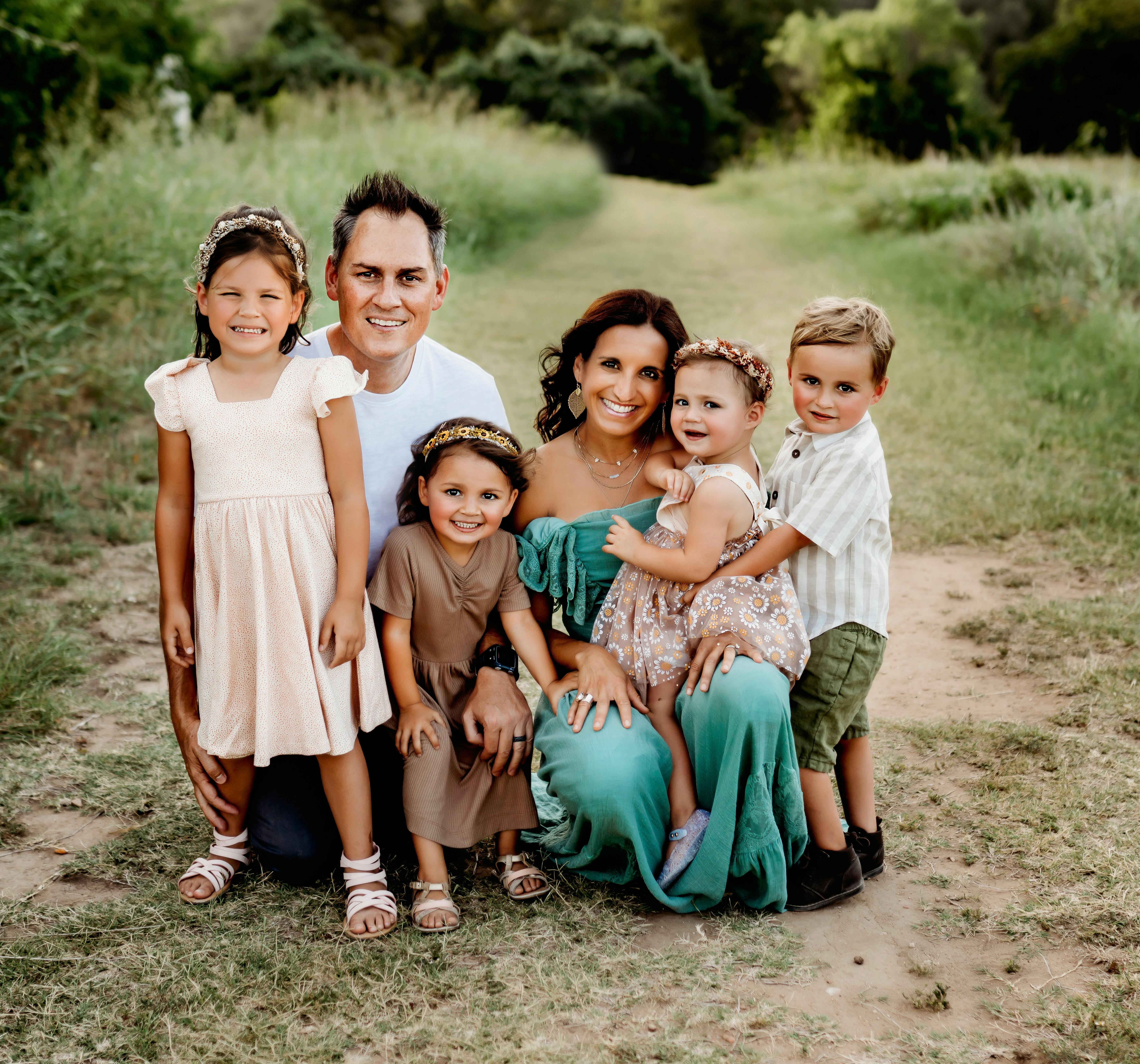 A cheerful family of six poses on a scenic dirt road surrounded by lush greenery.