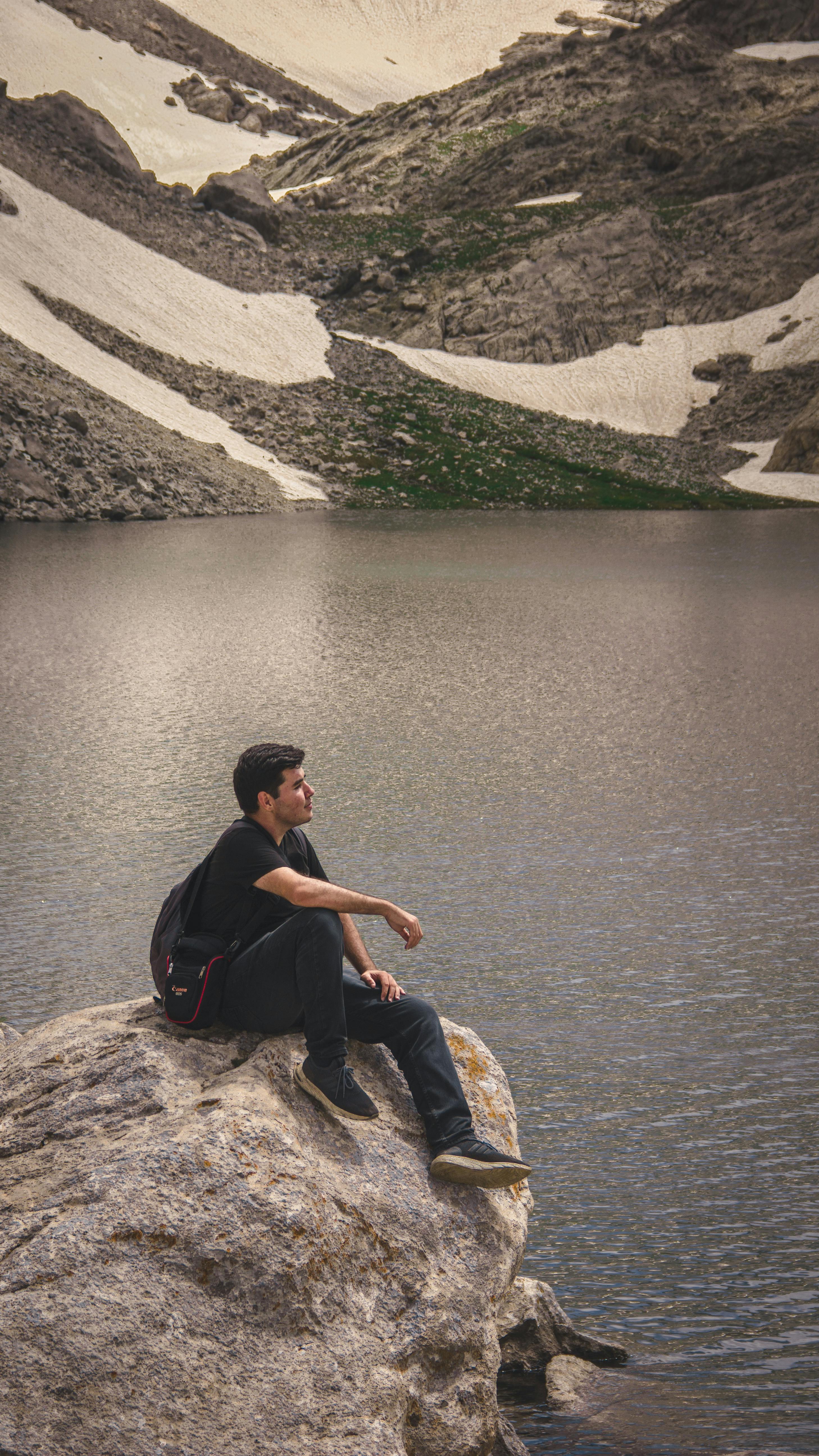 Prescription Goggle Inserts - A man sitting on a rock by a calm mountain lake, surrounded by snow-capped peaks.