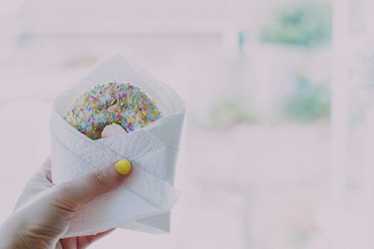 A close-up of a hand with yellow nails holding a sprinkle-covered donut wrapped in a napkin.