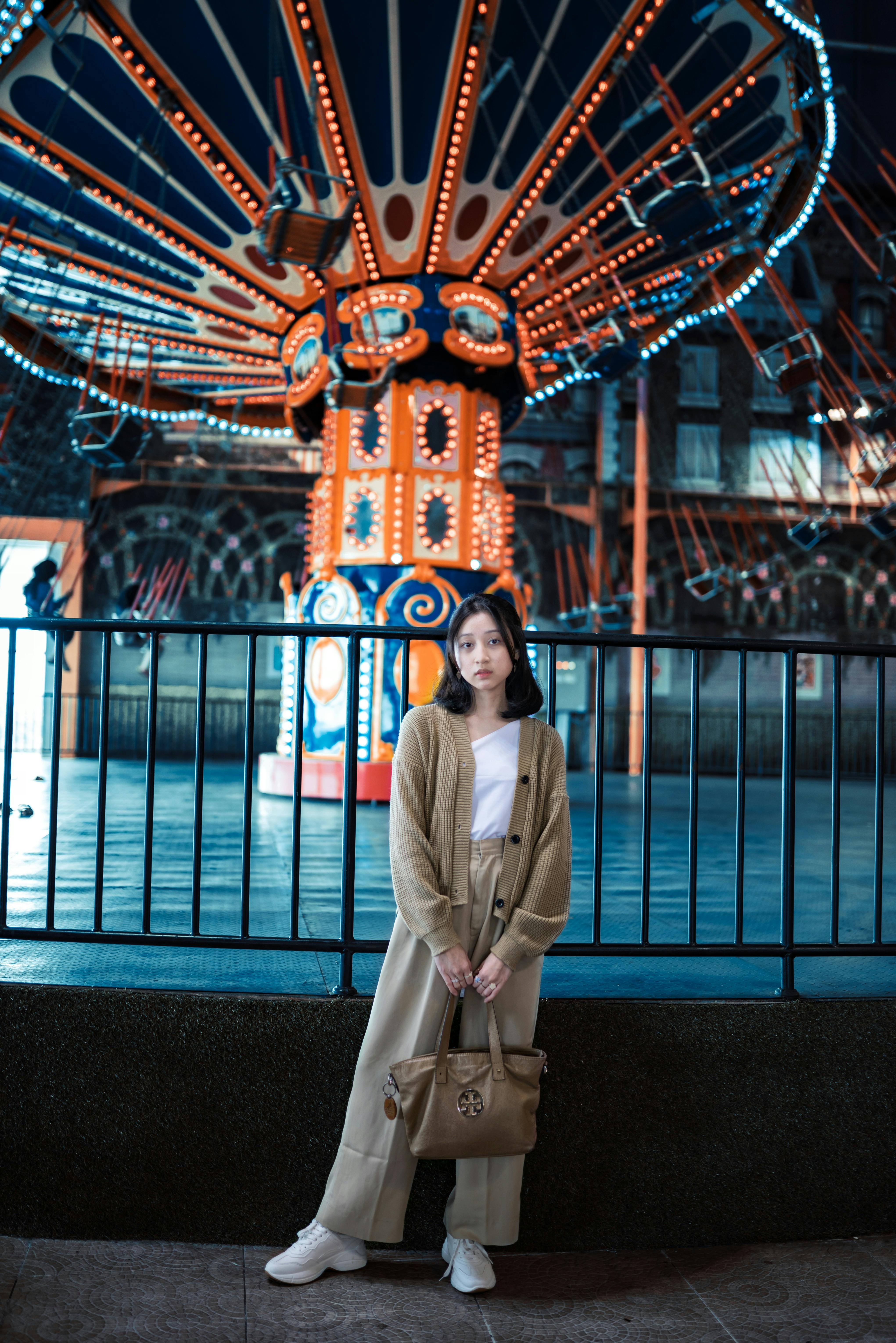 Free Asian woman in beige outfit stands stylishly in front of illuminated amusement ride. Stock Photo