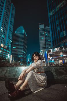 Stylish woman in dress posing at night in a vibrant city skyline.