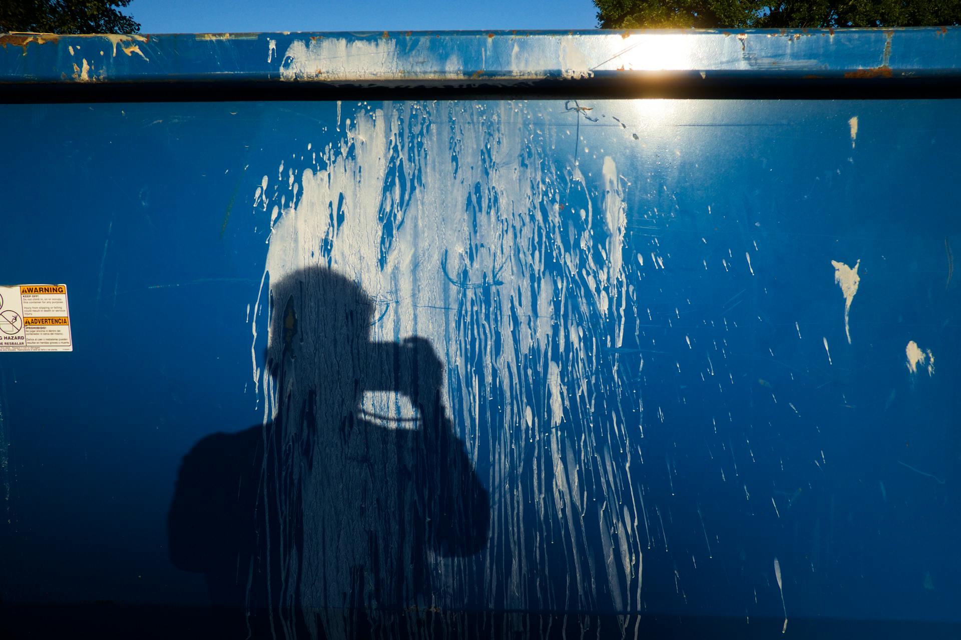 Shadow of a photographer on a blue trash container with stains and sunlight.