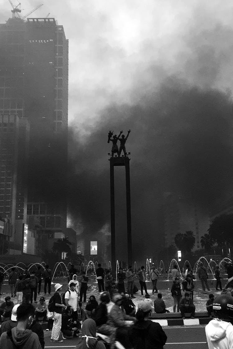 Crowd Gathered By A City Fountain, And Smoke In Background