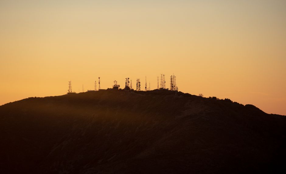 Communication towers silhouetted against an orange sunset sky on a hill.