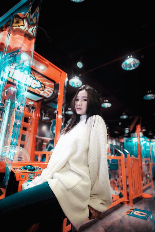 Free Fashionable Asian woman in a white sweater posing in a vibrant arcade. Modern and lively atmosphere. Stock Photo