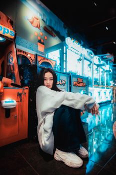Asian woman sitting in an arcade surrounded by game machines, wearing a white sweater.