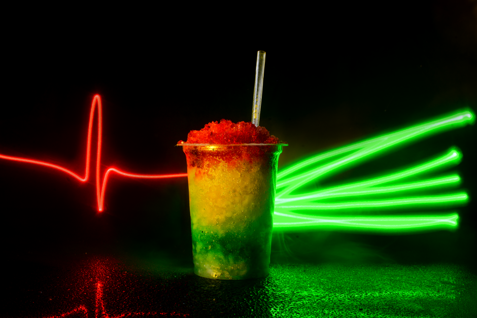 A paper snow cone cup filled with coarse shaved ice soaked in bright red and blue syrup, with a straw and spoon on the side.