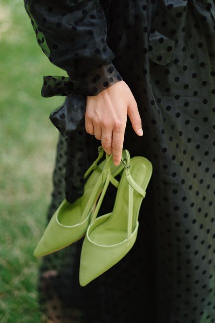 A close-up of a hand holding stylish green high heel shoes against a textured black dress.