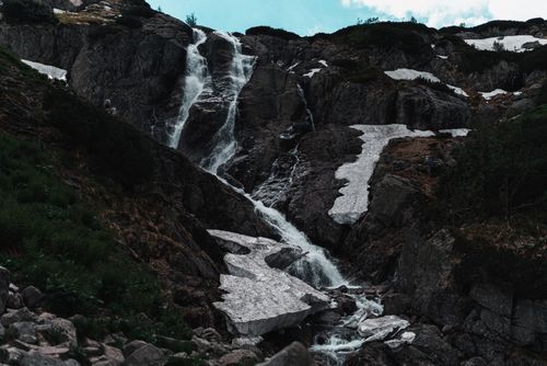 Free Majestic waterfall flows over rugged rocks amidst snow and lush greenery. Stock Photo