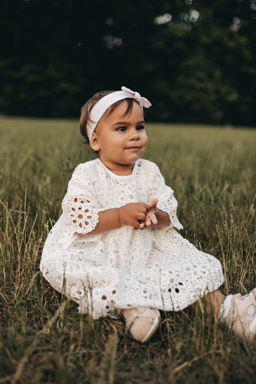 A Cute Toddler in White Dress and Headband Sitting on a Grass