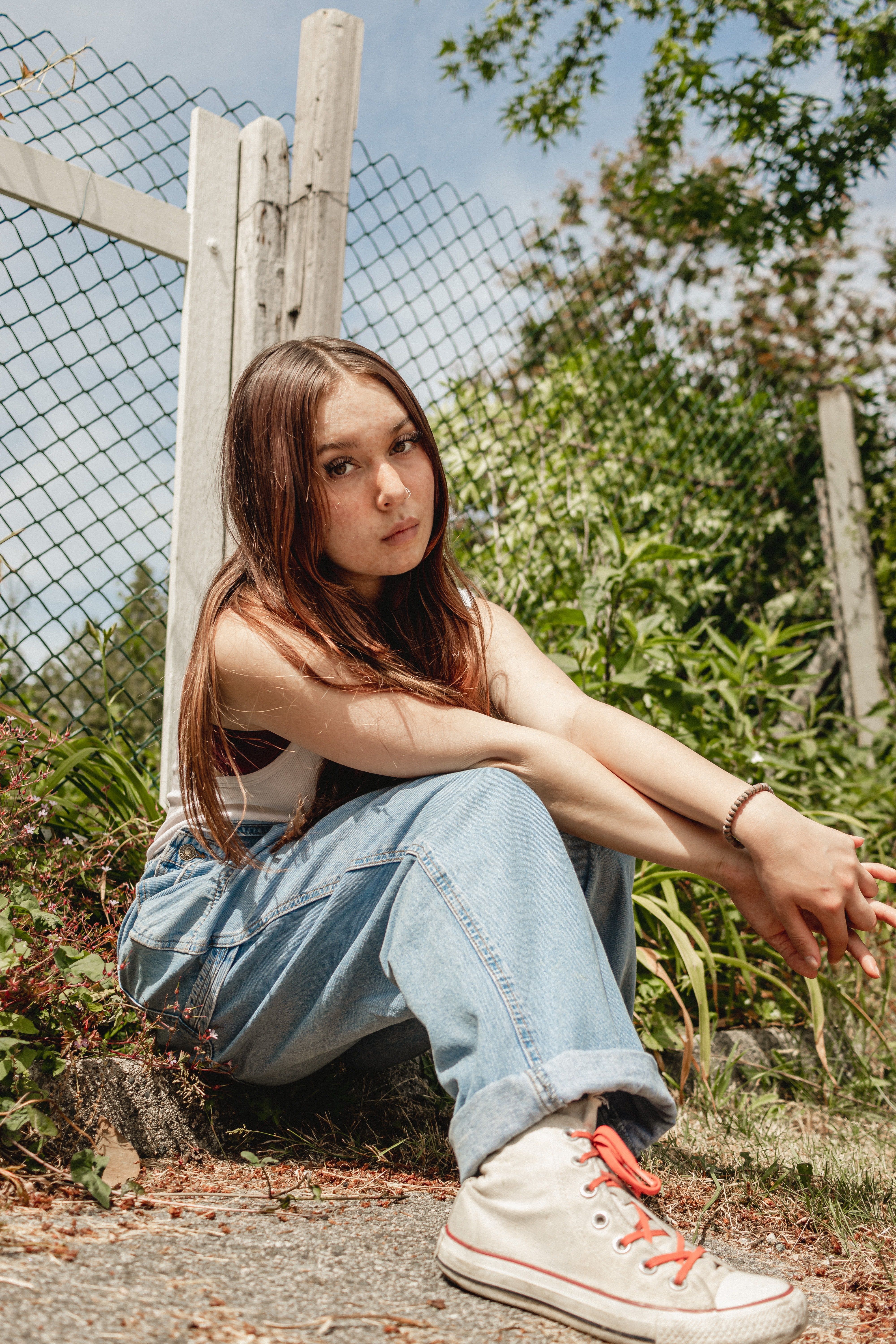 A young woman sitting outdoors in casual attire near a fence, exuding a relaxed vibe.