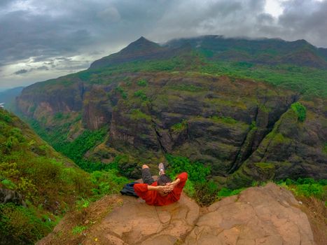 A person in a red jacket relaxes on a mountain top with a breathtaking view of cliffs and lush greenery.