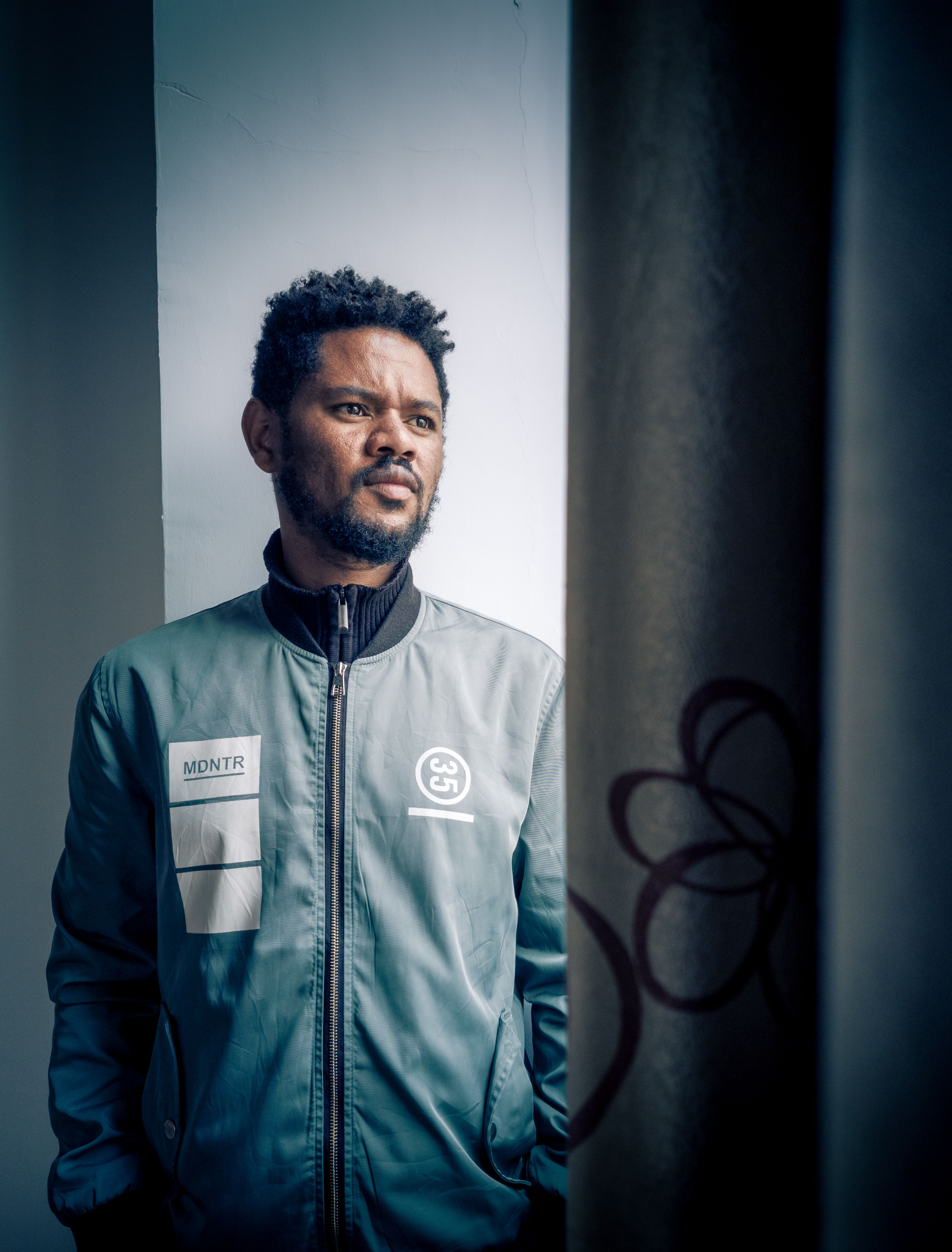 A thoughtful man stands by a window, wearing a casual jacket, looking into the distance.