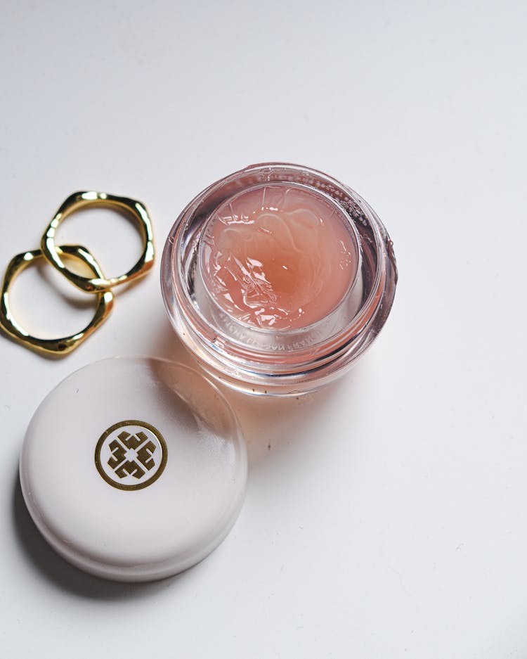 Gold Rings And Cosmetic Gel In A Jar Against White Background