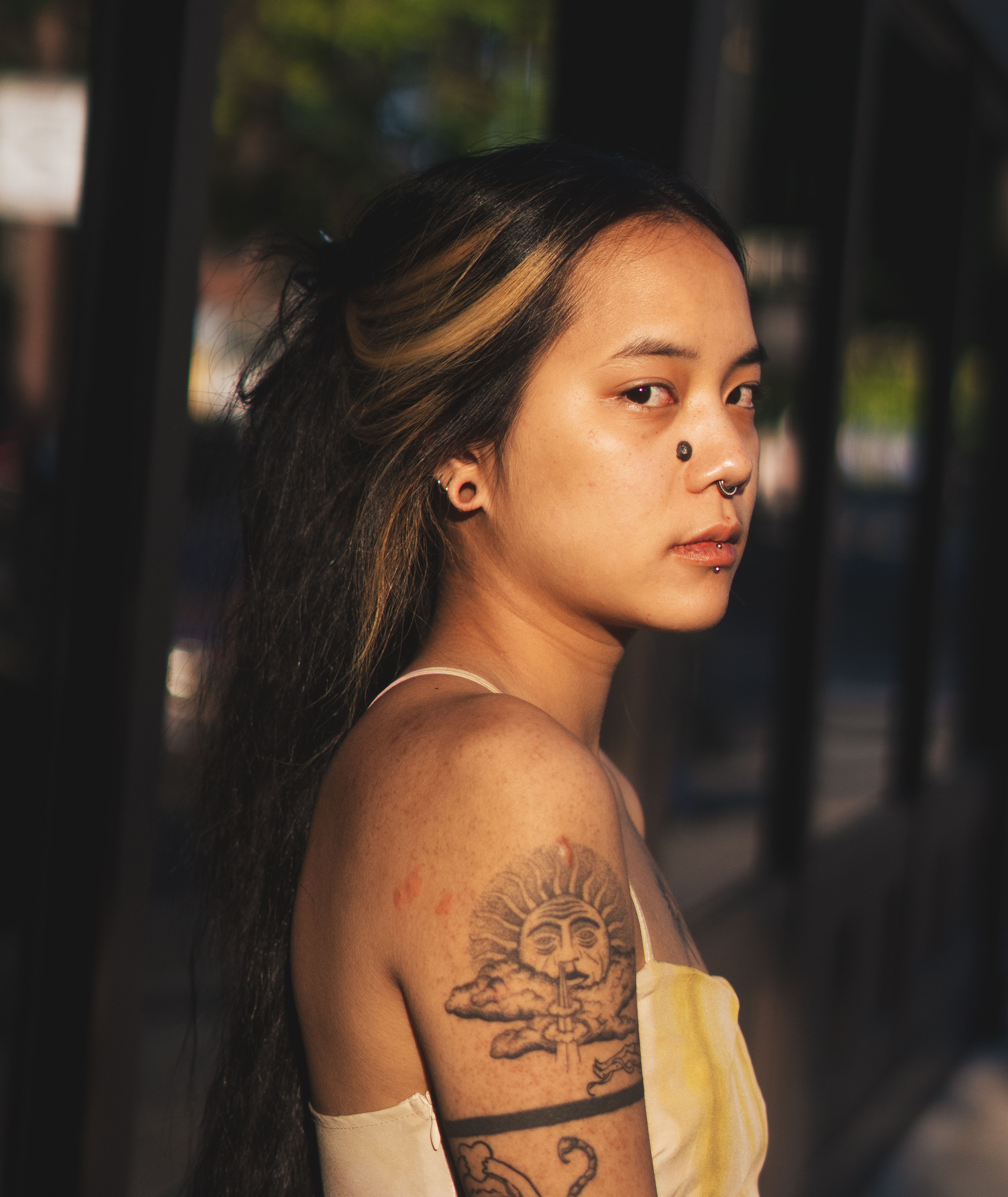 A young woman with tattoos stands outdoors, displaying a confident, contemplative expression.