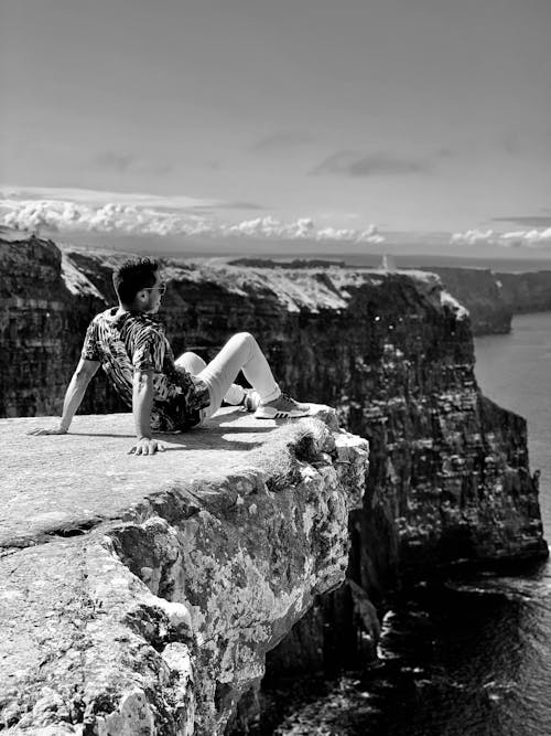 A Grayscale of a Man Sitting on a Cliff