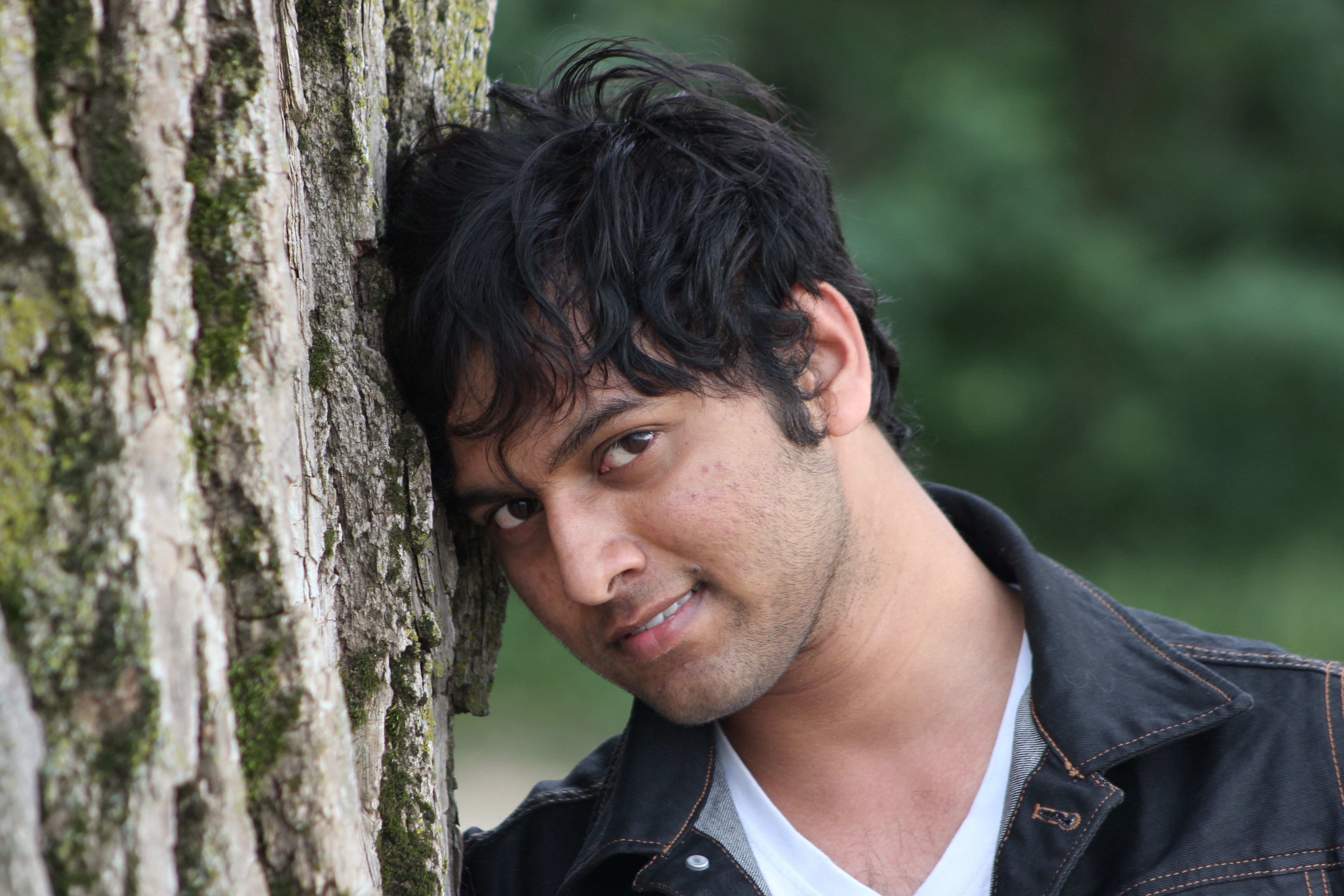 Portrait of a young man leaning on a mossy tree with a playful expression in an outdoor setting.