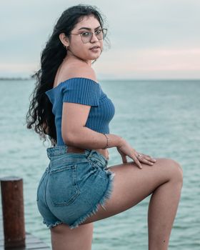 Stylish woman posing by the ocean in a blue top and denim shorts, showcasing fashion and confidence.