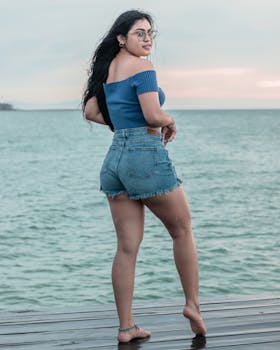 Woman standing on a wooden deck by the ocean wearing denim shorts and an off-shoulder top.