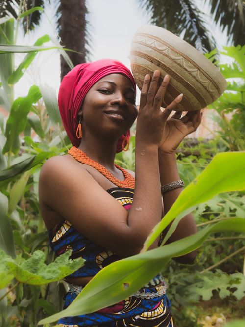 Free Smiling woman in vibrant attire holding a bowl amidst lush Nigerian garden. Stock Photo