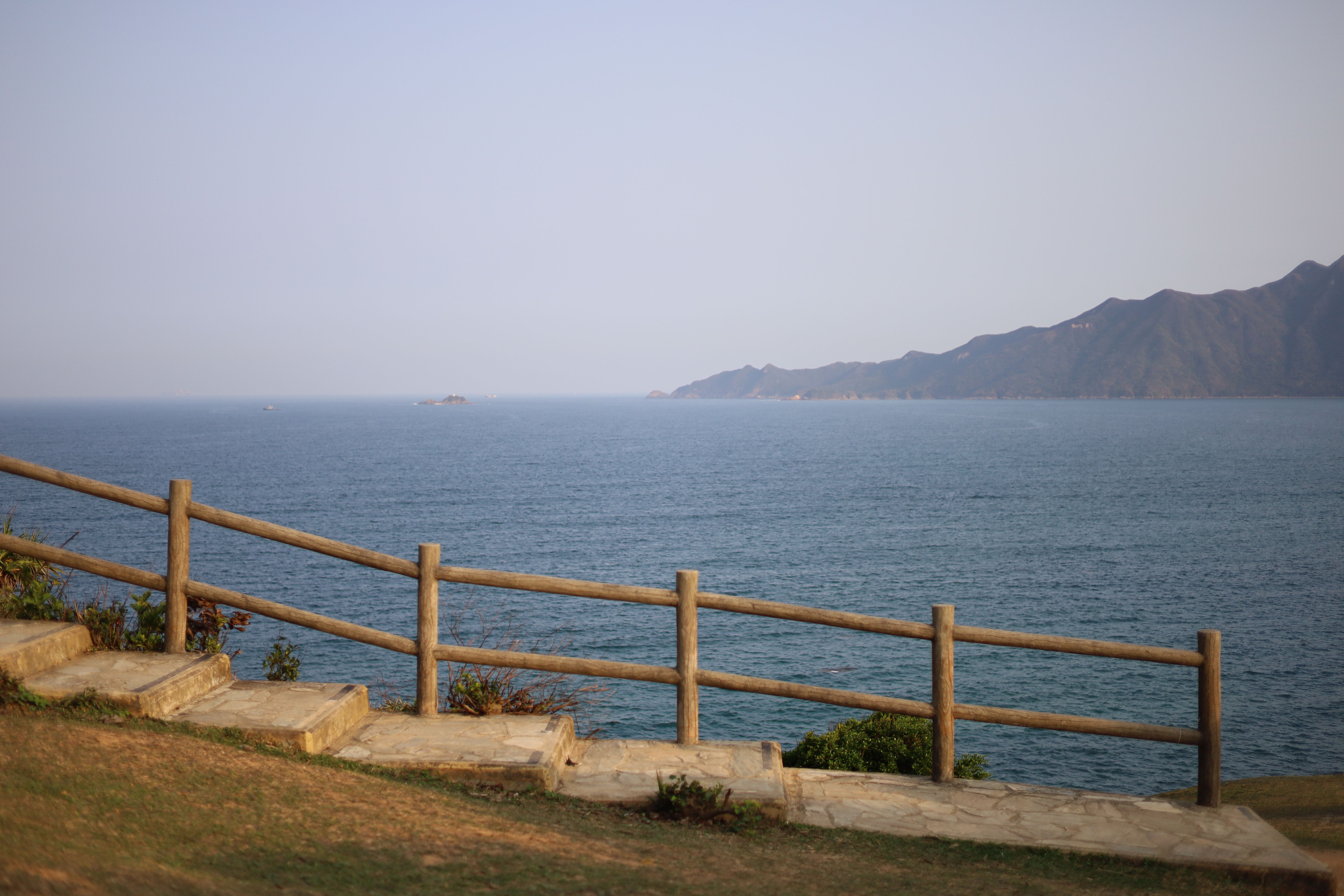 Fotomural Camino costero tranquilo con barandilla de madera que ofrece vistas panorámicas del océano y las montañas bajo cielos azules claros.