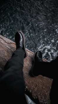 Black shoes and trousers capture a man's legs sitting on a bridge over rushing river.