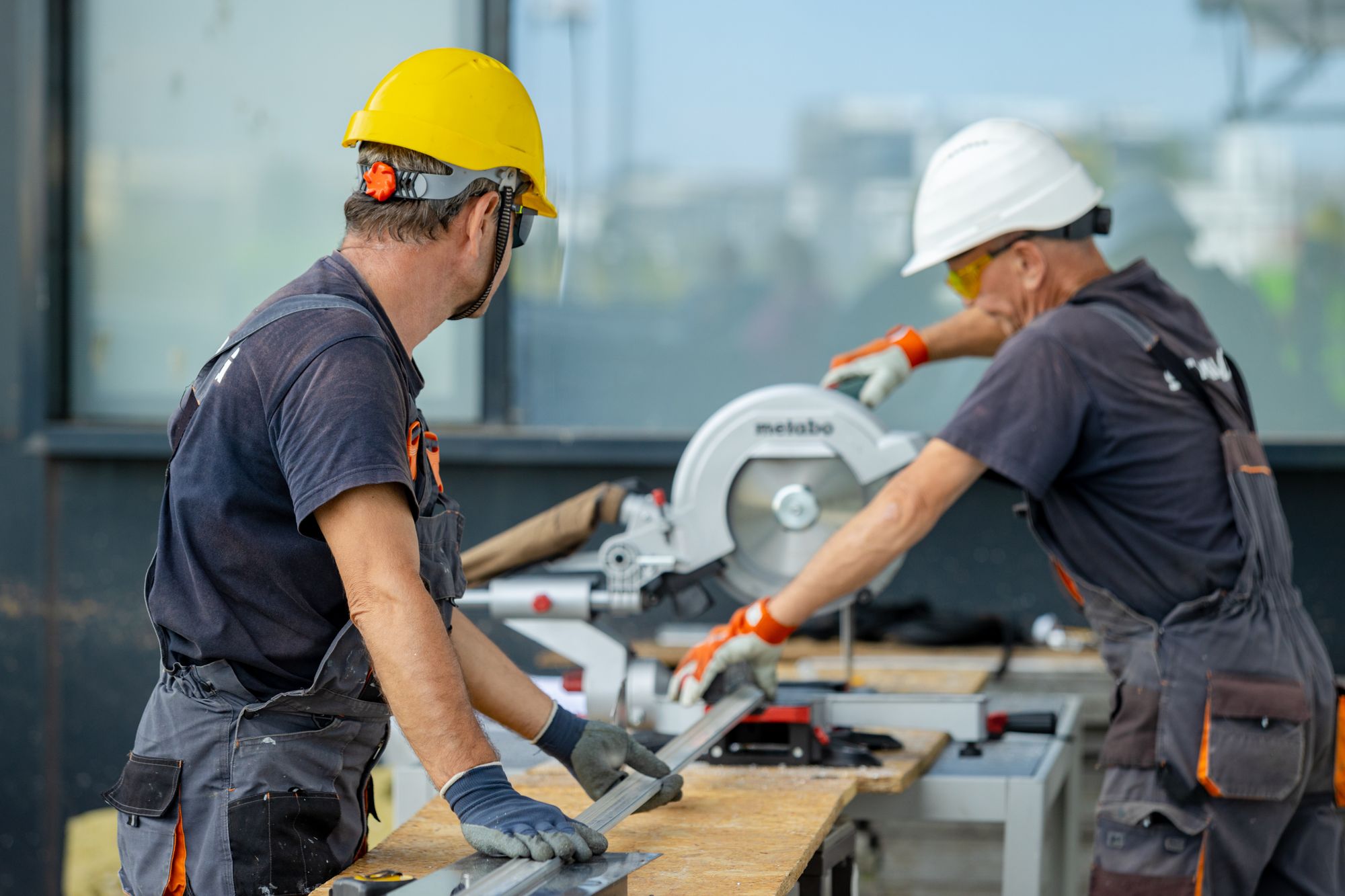 Two tradespeople wearing safety equipment and working with a mitre saw