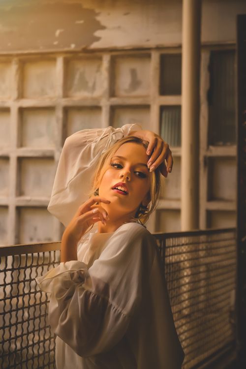 Free Caucasian woman with blonde hair posing gracefully by a metal railing in warm ambient light. Stock Photo