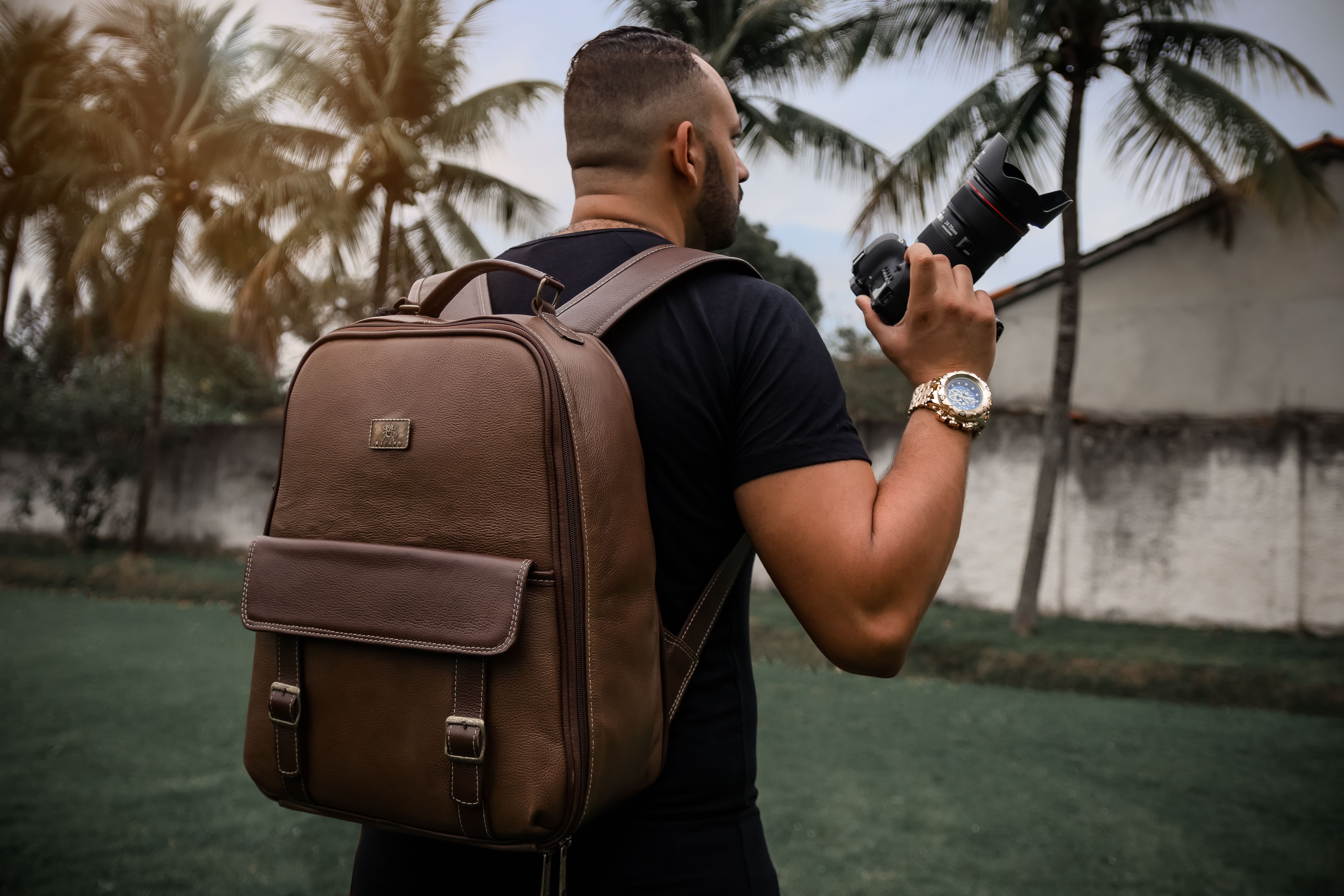 Adult male stands outdoors holding camera with backpack in tropical Rio de Janeiro.