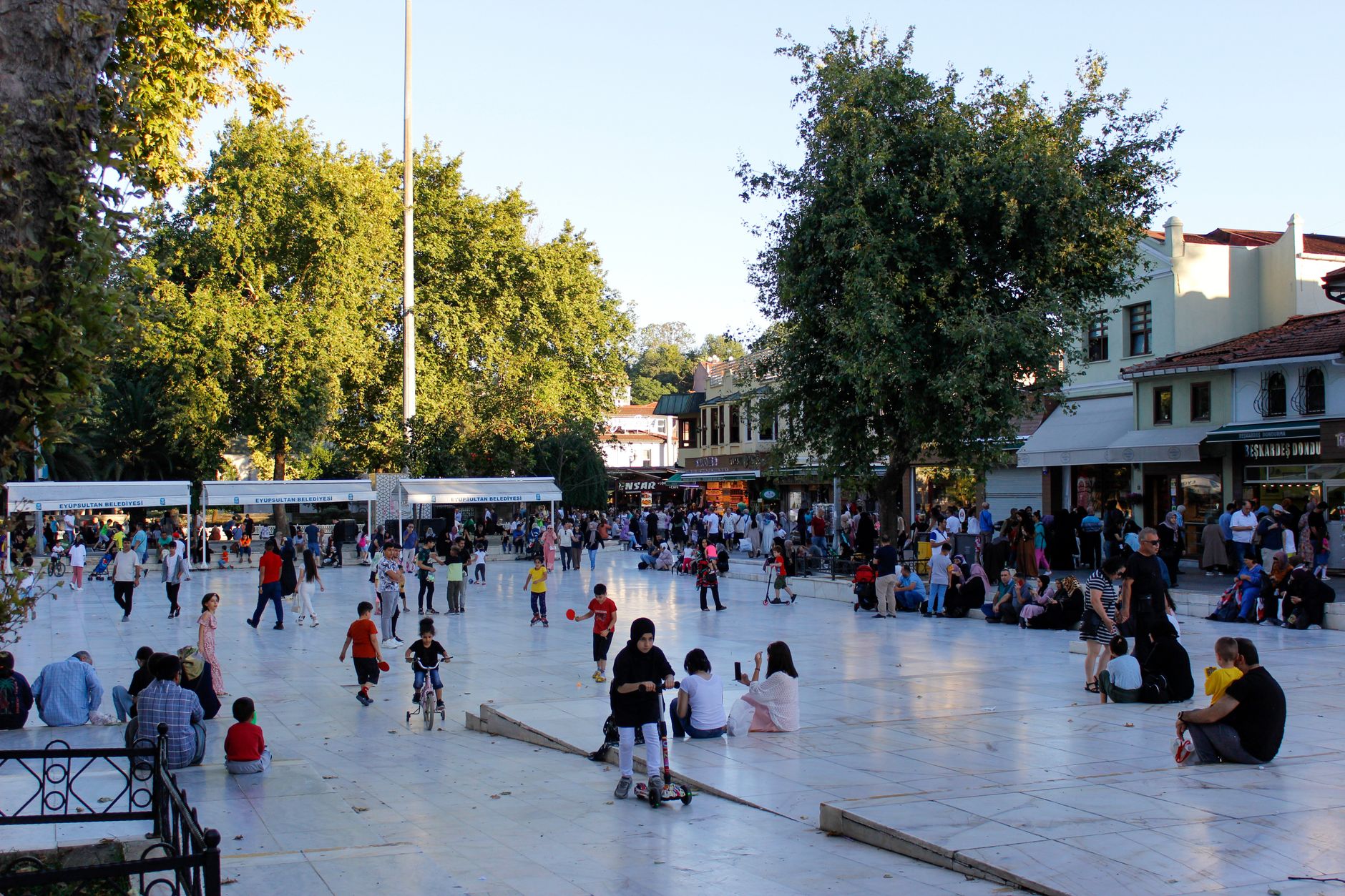 台中親子景點推薦:爸媽帶兩個以上小孩也handle得住的7大去處 A vibrant city square filled with families, children playing and relaxing under green trees on a sunny day.