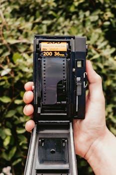 A hand holding an open film camera with a visible 35mm film roll against a leafy background.