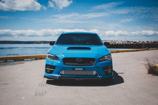 A vibrant blue sports car parked near the ocean, under a clear blue sky.