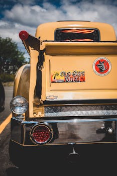 Close-up of a vintage yellow Chevrolet with retro details and a humorous bumper sticker under sunny skies.
