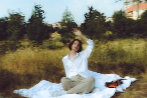 Free Artistic image of a woman sitting on a white blanket in a grassy field with trees in the background, featuring a blurred effect. Stock Photo