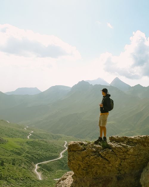 Free A lone hiker on a cliff, overlooking a vast mountain landscape under a bright sky. Stock Photo
