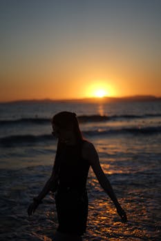A silhouette of a woman walking by the beach at sunset, capturing the calm ocean view and warm hues.