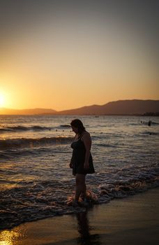 A woman enjoys a serene walk along the beach at sunset, with golden skies and gentle waves.