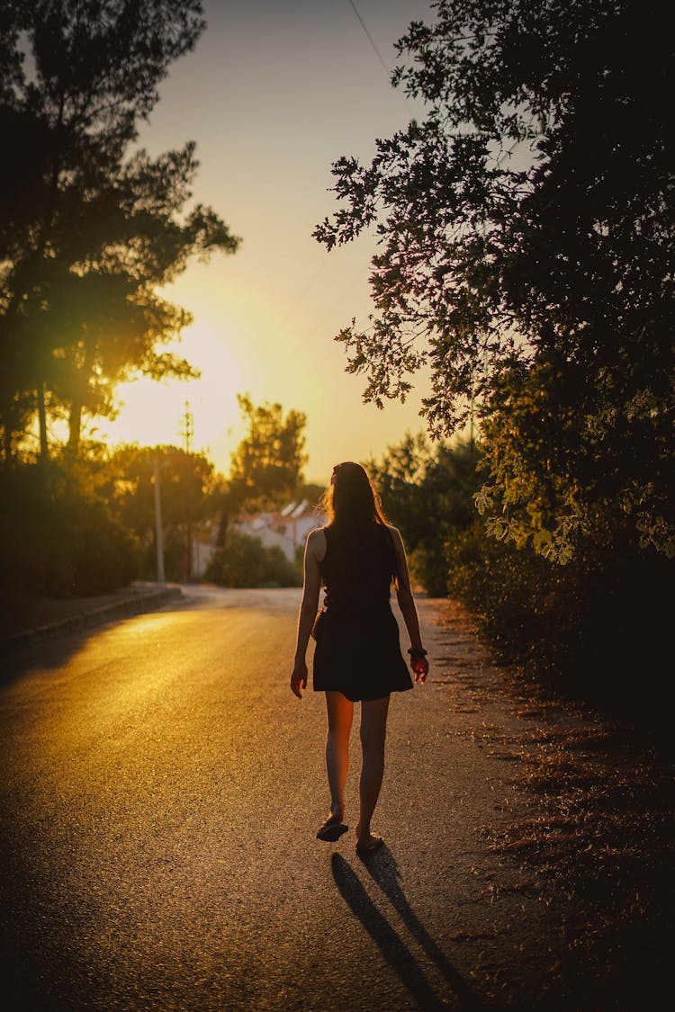 Back View Of A Woman In Black Dress Walking On The Concrete Road During Sunset