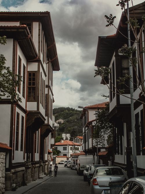 Free A picturesque street scene featuring traditional architecture, parked cars, and people walking under a cloudy sky. Stock Photo