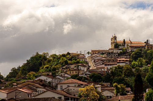 Free A picturesque view of Ouro Preto's historic hilltop church surrounded by lush greenery under dramatic skies. Stock Photo