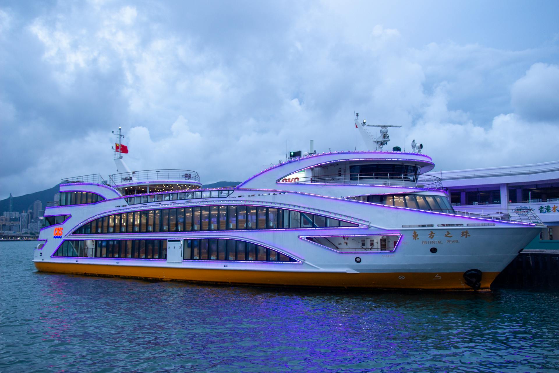 A luxurious cruise ship docked by the port, illuminated with vibrant lights against a cloudy twilight sky.