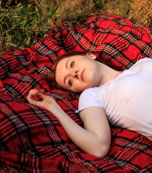 Free A woman relaxing on a red plaid blanket in a grassy outdoor setting, enjoying a tranquil moment. Stock Photo