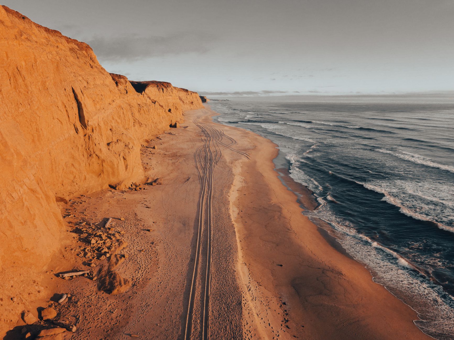 Aerial view of Half Moon Bay coastline with rugged cliffs and tire tracks on the sandy shore.