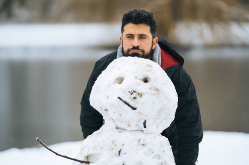 Free A bearded man poses with a snowman beside a lake during winter. Stock Photo