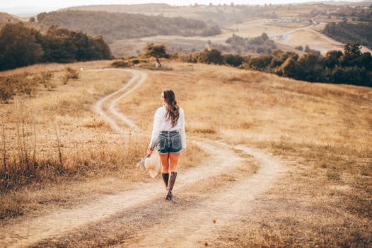 A woman in shorts and boots walks on a winding rural path with a scenic countryside view.